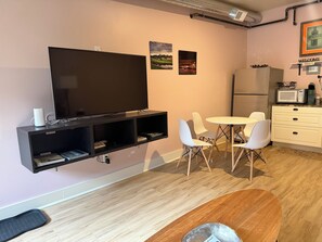 A modern living space with a flat-screen TV mounted on the wall, a dining area with a round table and four chairs, a kitchen area with a microwave and refrigerator, and a small shelf with books. - Walk to everything! This apartment puts you in the heart of historic Galena! (Galena)