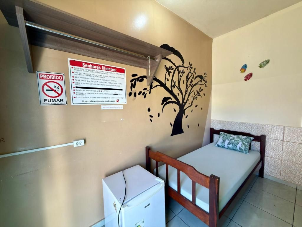 Quarto de hotel com cama, pequeno frigorífico branco, prateleira fixada na parede e um decalque de árvore na parede.