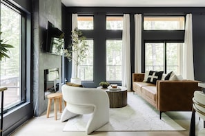 A modern living room with a sofa, armchair, and coffee table, large windows with curtains, and a fireplace. - The Den at Dunlap Ridge (Laurel Township)