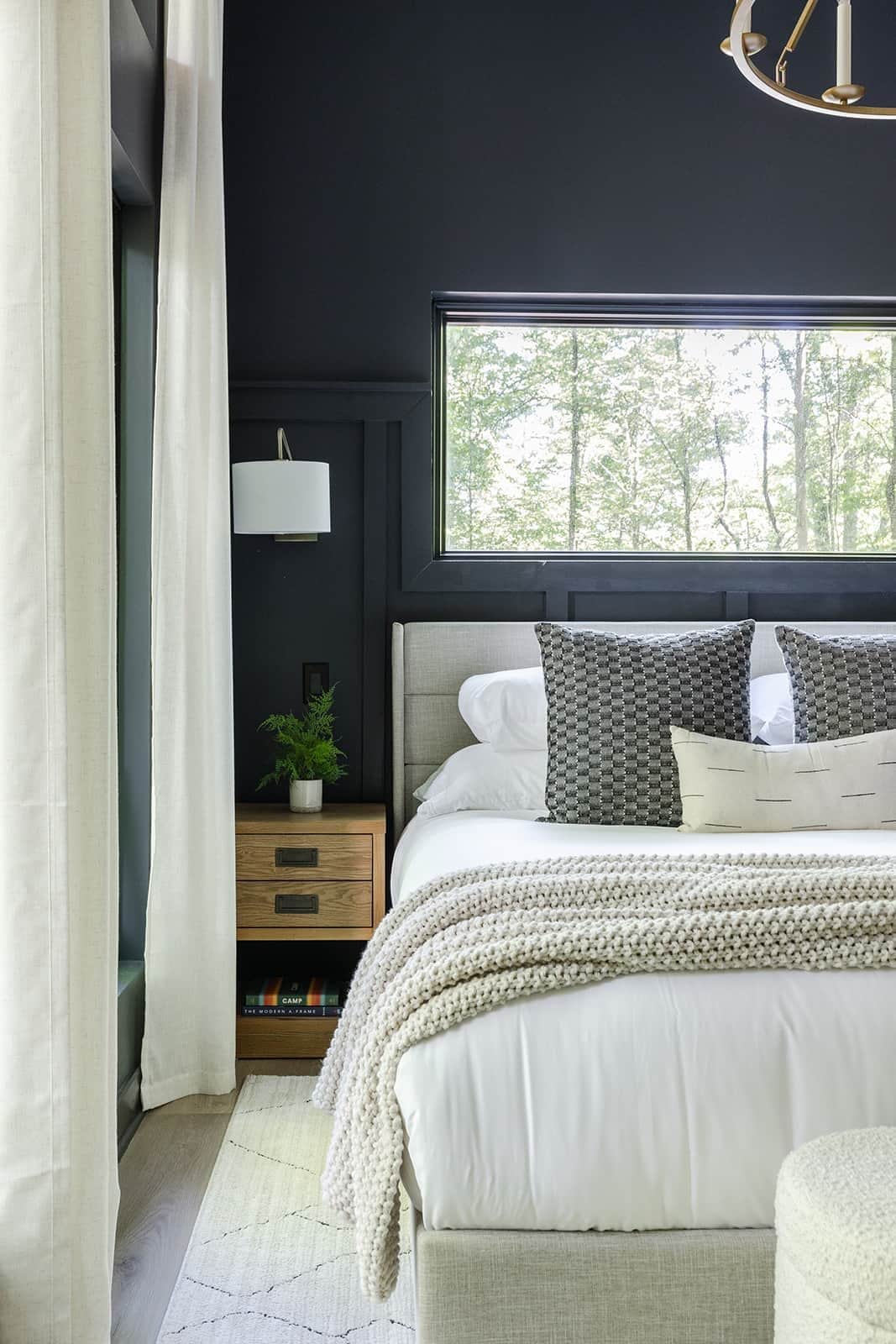 Une chambre avec une grande fenêtre, un lit avec de nombreux oreillers, une table de chevet avec une plante et une lampe fixée au mur.