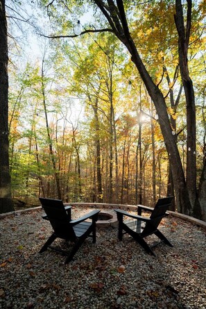 An outdoor seating area with a fire pit and benches, surrounded by trees with autumn foliage. - The Pine at Dunlap Ridge (Laurel Township)