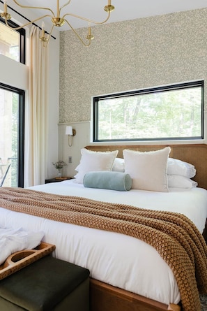 A hotel room with a large bed, a wooden headboard, a window with a view of trees, and a chandelier. - The Pine at Dunlap Ridge (Laurel Township)