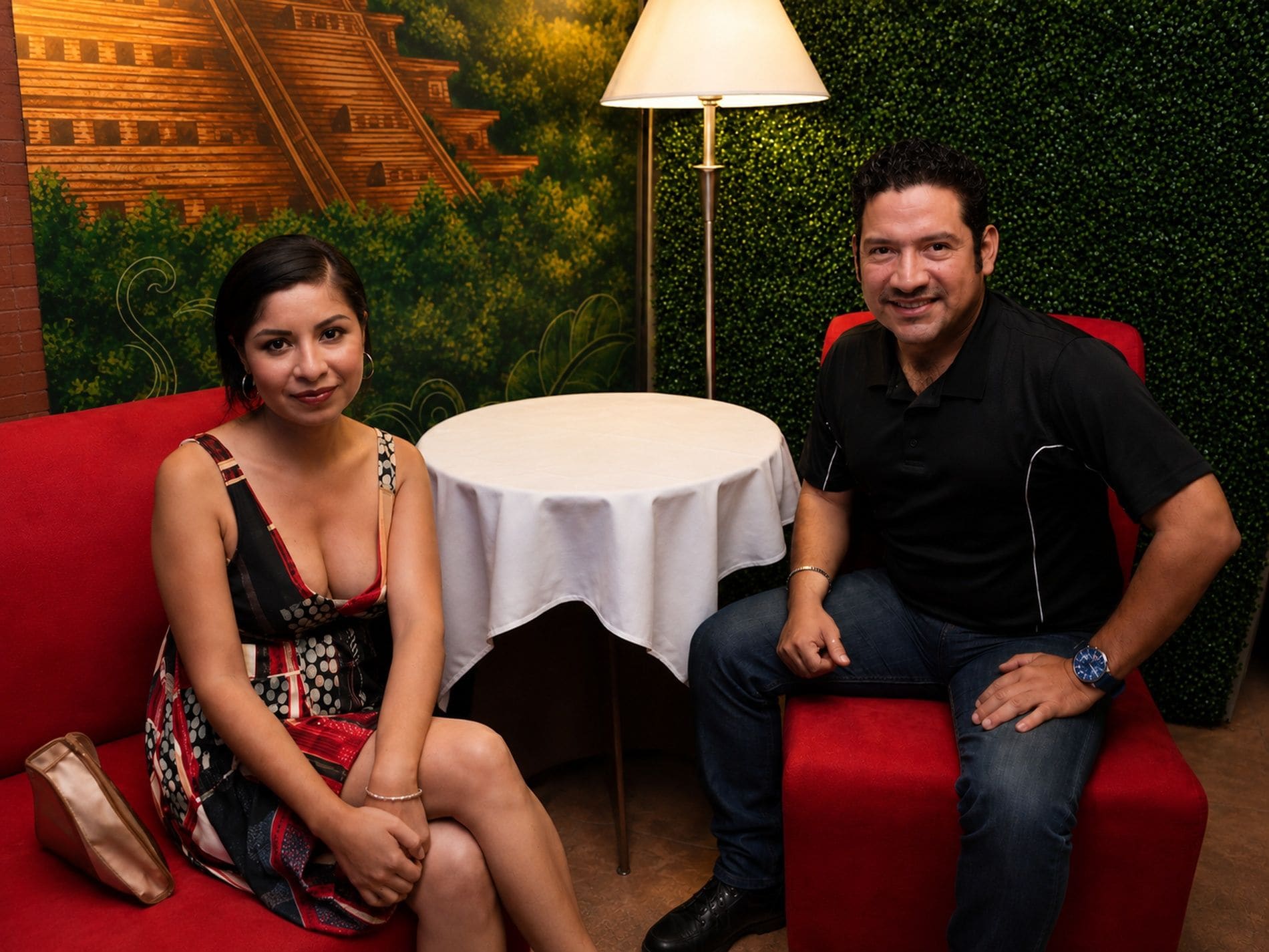 Dos personas sentadas en sofás rojos con una pequeña mesa redonda entre ellas, una lámpara y una pared con un diseño de hojas verdes.