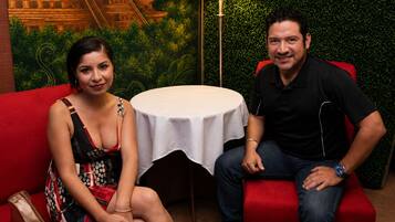Dos personas sentadas en sofás rojos con una pequeña mesa redonda entre ellas, una lámpara y una pared con un diseño de hojas verdes.