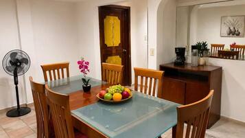 A dining area with a glass-top table, wooden chairs, a fruit bowl, a fan, and a kitchen area with a coffee maker.