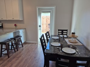 A dining area with a table set for four, wooden chairs, a small side table with a plant, and a view of a wooden fence through an open door.