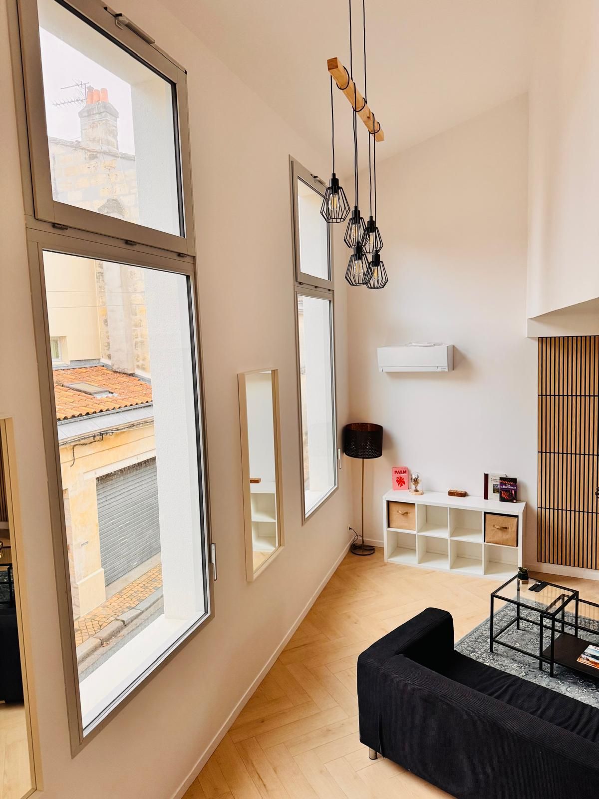 A modern living room with large windows, a black sofa, a wooden floor, and a white console table with decorative items.