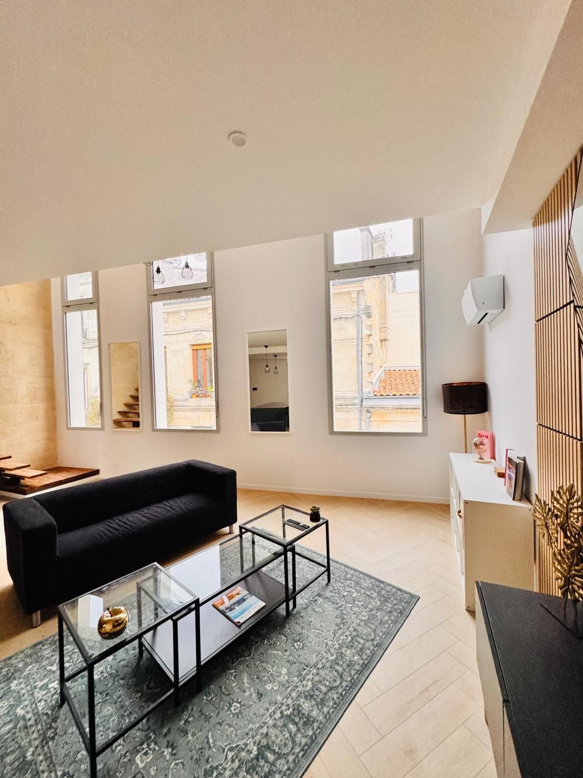 A modern living room with a black sofa, glass coffee table, and large windows offering views of buildings.