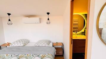 A bedroom with a bed, bedside tables, an air conditioning unit, and a view into a bathroom.
