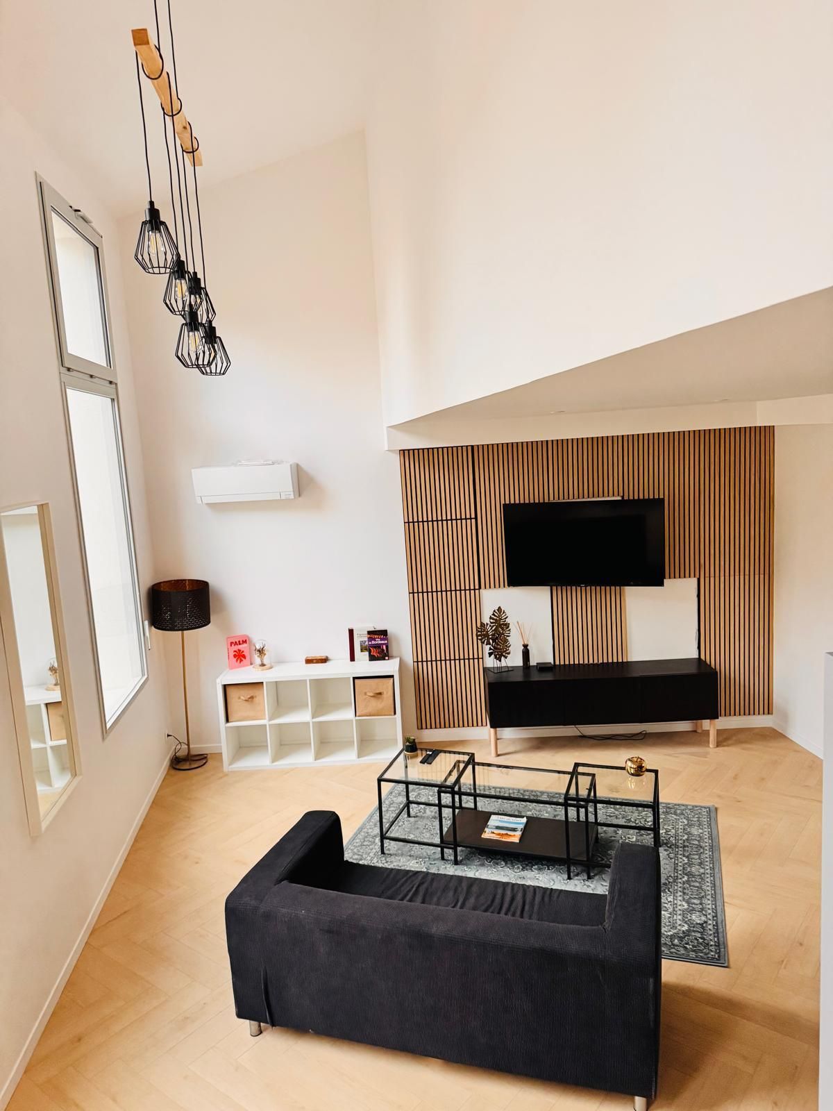 A modern living room with a black sofa, a glass coffee table, a TV mounted on a wooden panel wall, and a floor lamp.