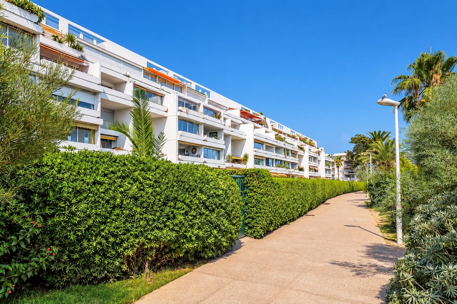 A modern multi-story hotel with balconies, a paved walkway, and lush greenery.
