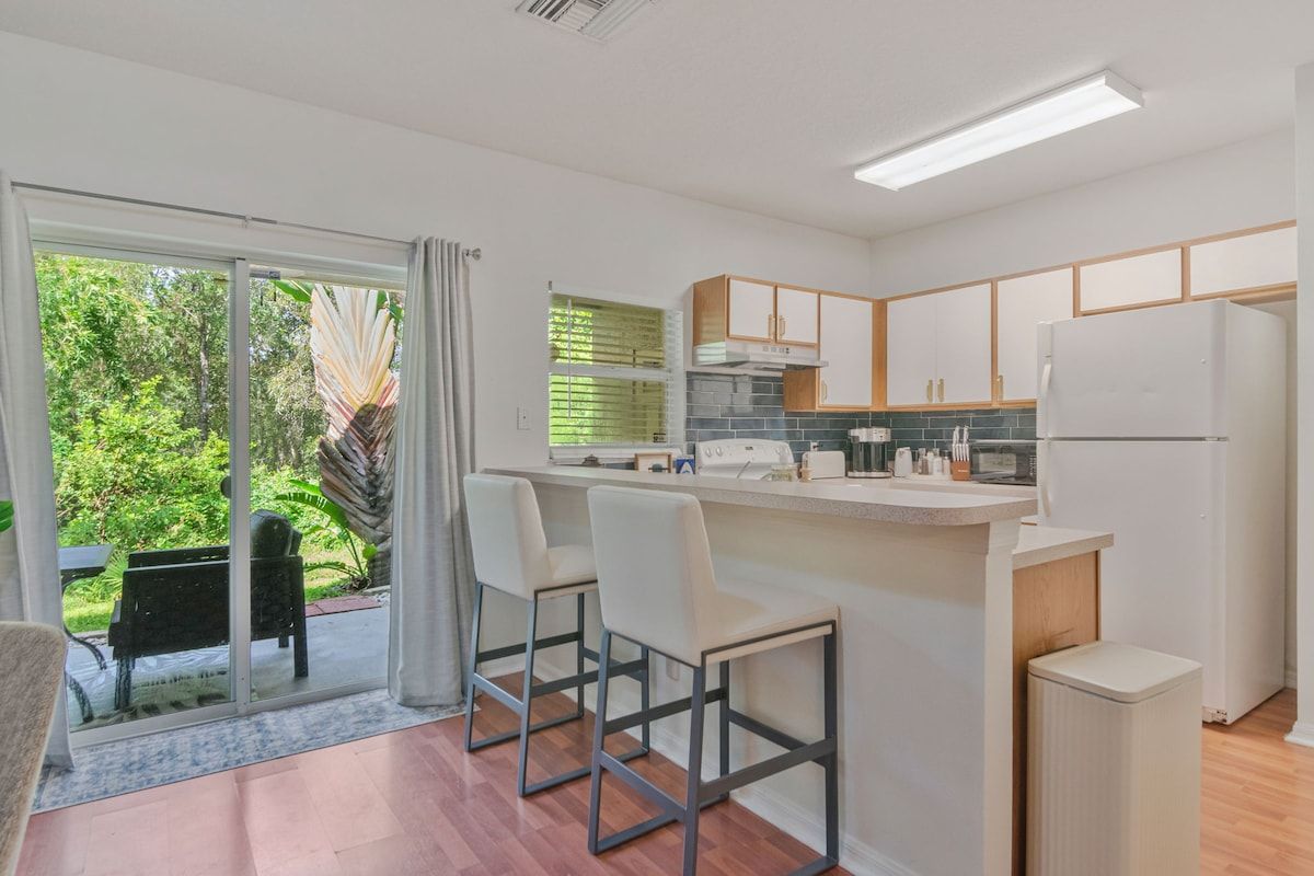 A modern kitchen with a central island, bar stools, and a view of greenery outside.