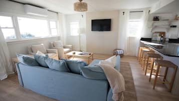 Una sala de estar moderna con un sofá azul, un televisor de pantalla plana y una zona de cocina con taburetes de bar.