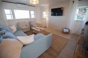 Un salon comprenant un canapé, des fauteuils, une table basse, un téléviseur et une fenêtre avec des rideaux.
