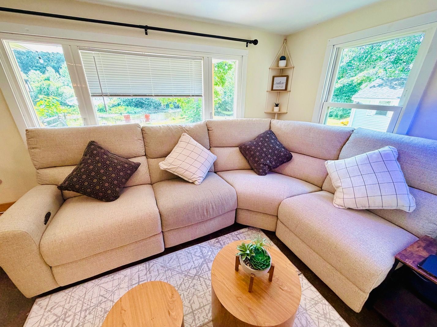 A beige sectional sofa with patterned cushions, a small wooden coffee table with a succulent plant, and a view of greenery through the windows.