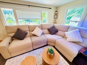 Un sofá seccional color beige con cojines estampados, una pequeña mesa de centro de madera con una suculenta, y vistas al verdor a través de las ventanas.