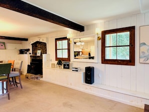 A room with a fireplace, a dining area with chairs, a kitchen with a window, and a wall-mounted speaker.