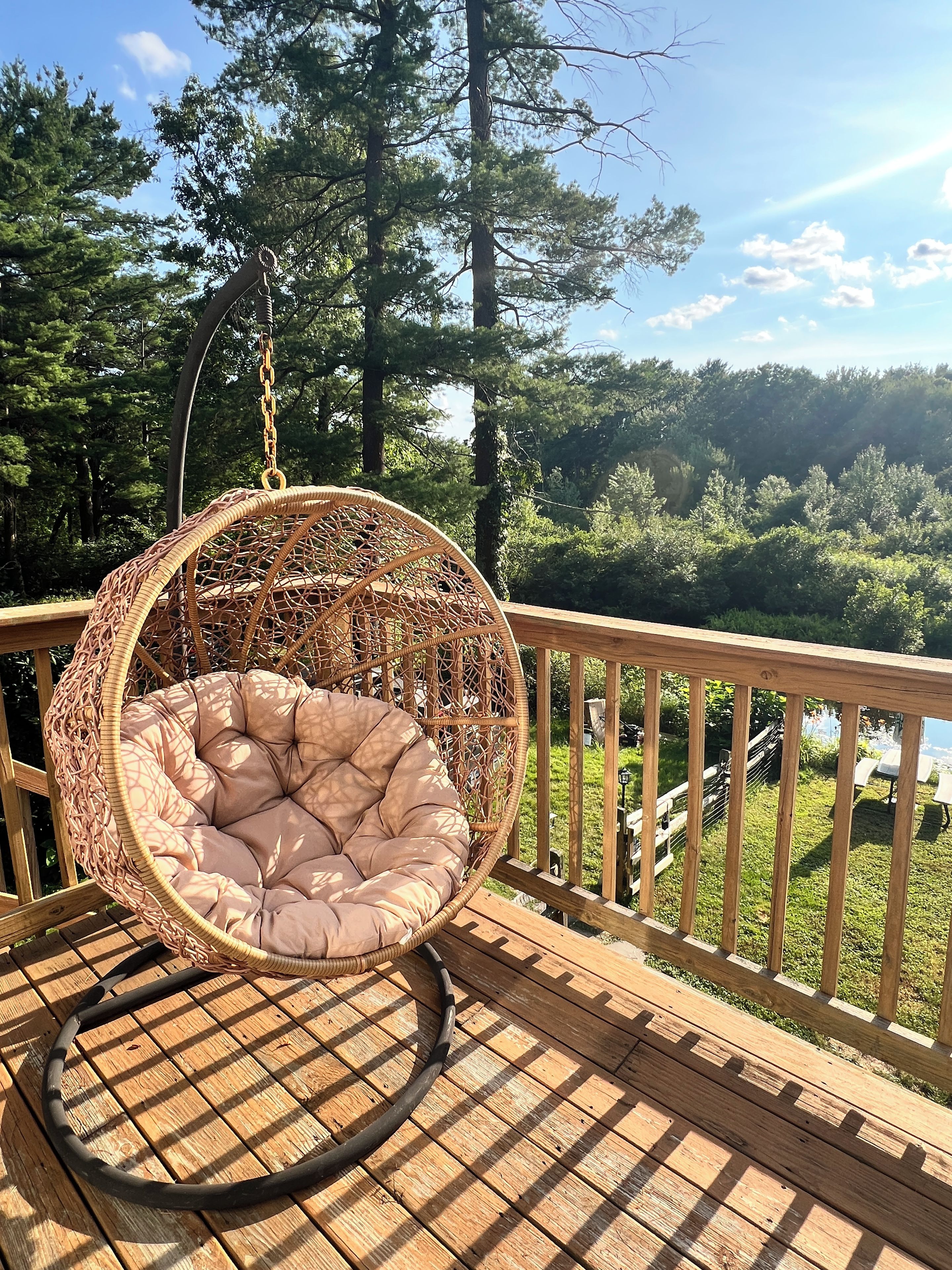 A wicker hanging chair with a cushioned seat on a wooden deck overlooking a forested area.