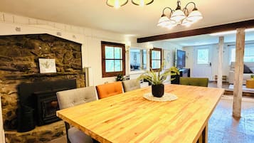 A spacious dining area with a wooden table, chairs, and a potted plant. A stone fireplace and a stone wall are visible in the background.