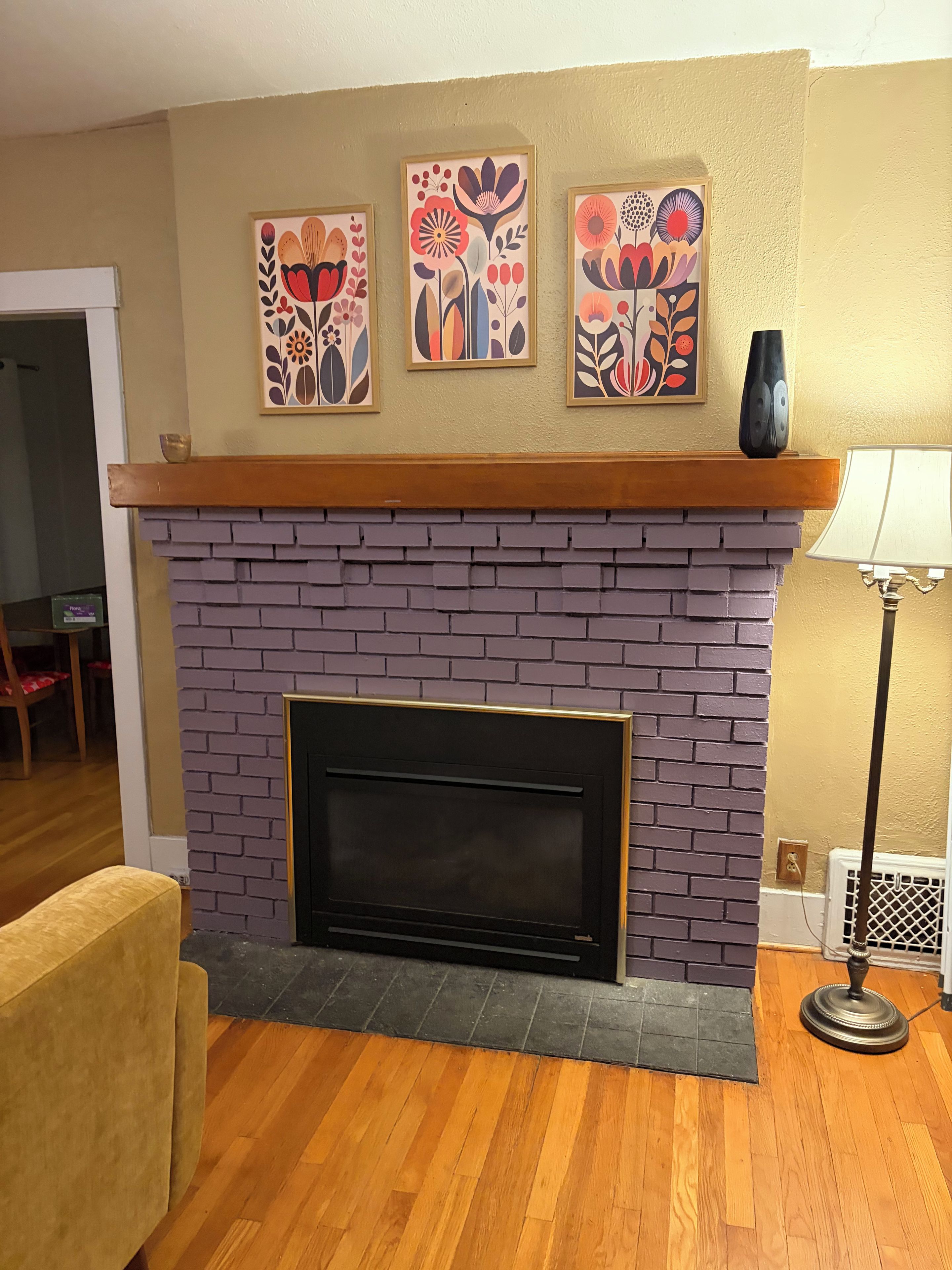 A living room with a brick fireplace, three framed floral artworks, a wooden mantel, and a yellow wall.