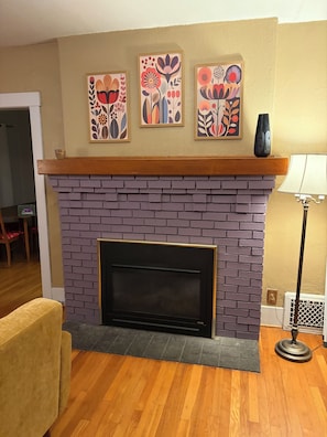 A living room with a brick fireplace, three framed floral artworks, a wooden mantel, and a yellow wall.