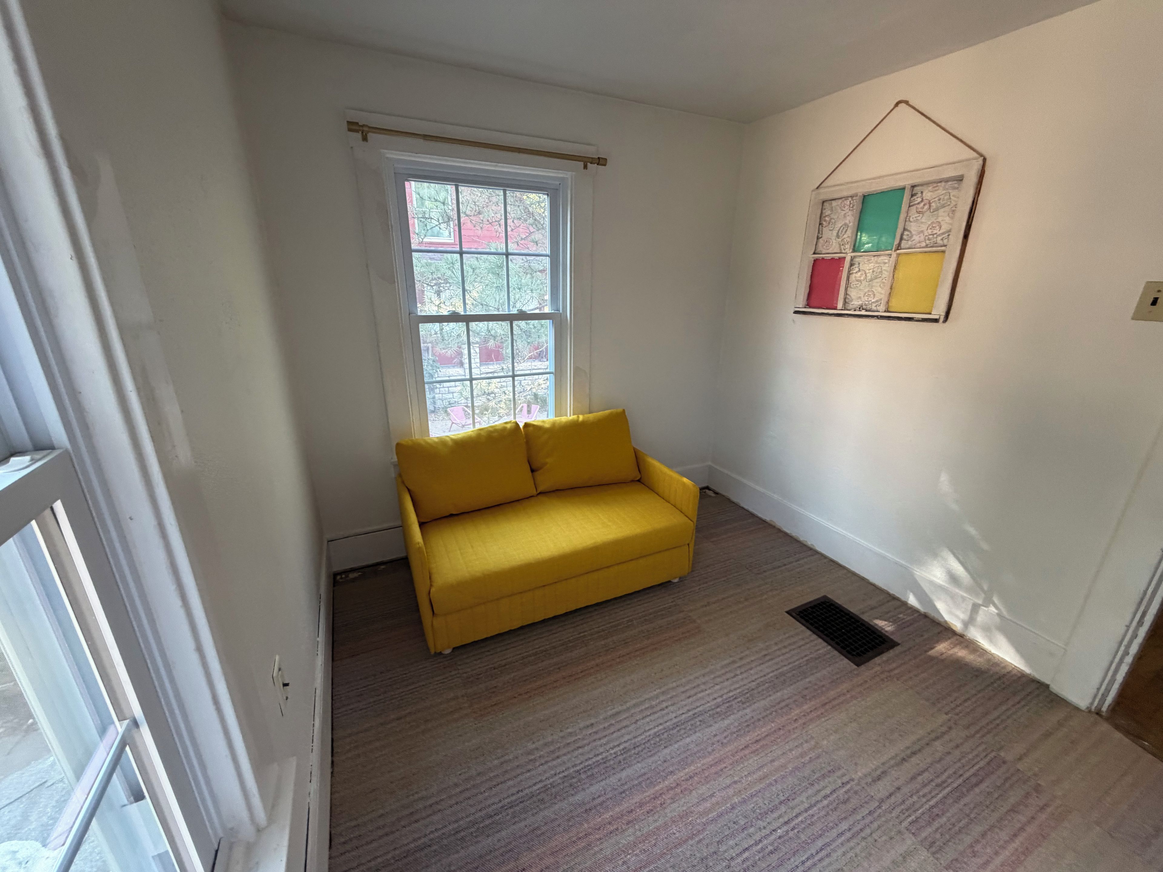 Ein Zimmer mit einem gelben Sofa, einem Fenster mit Blick auf Bäume und einem an der Wand hängenden Kunstwerk.