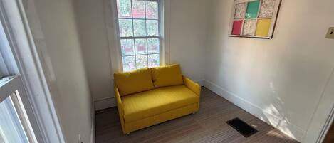A room with a yellow sofa, a window with a view of trees, and a wall-mounted artwork.