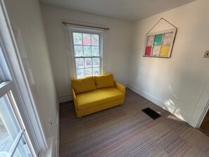 A room with a yellow sofa, a window with a view of trees, and a wall-mounted artwork.