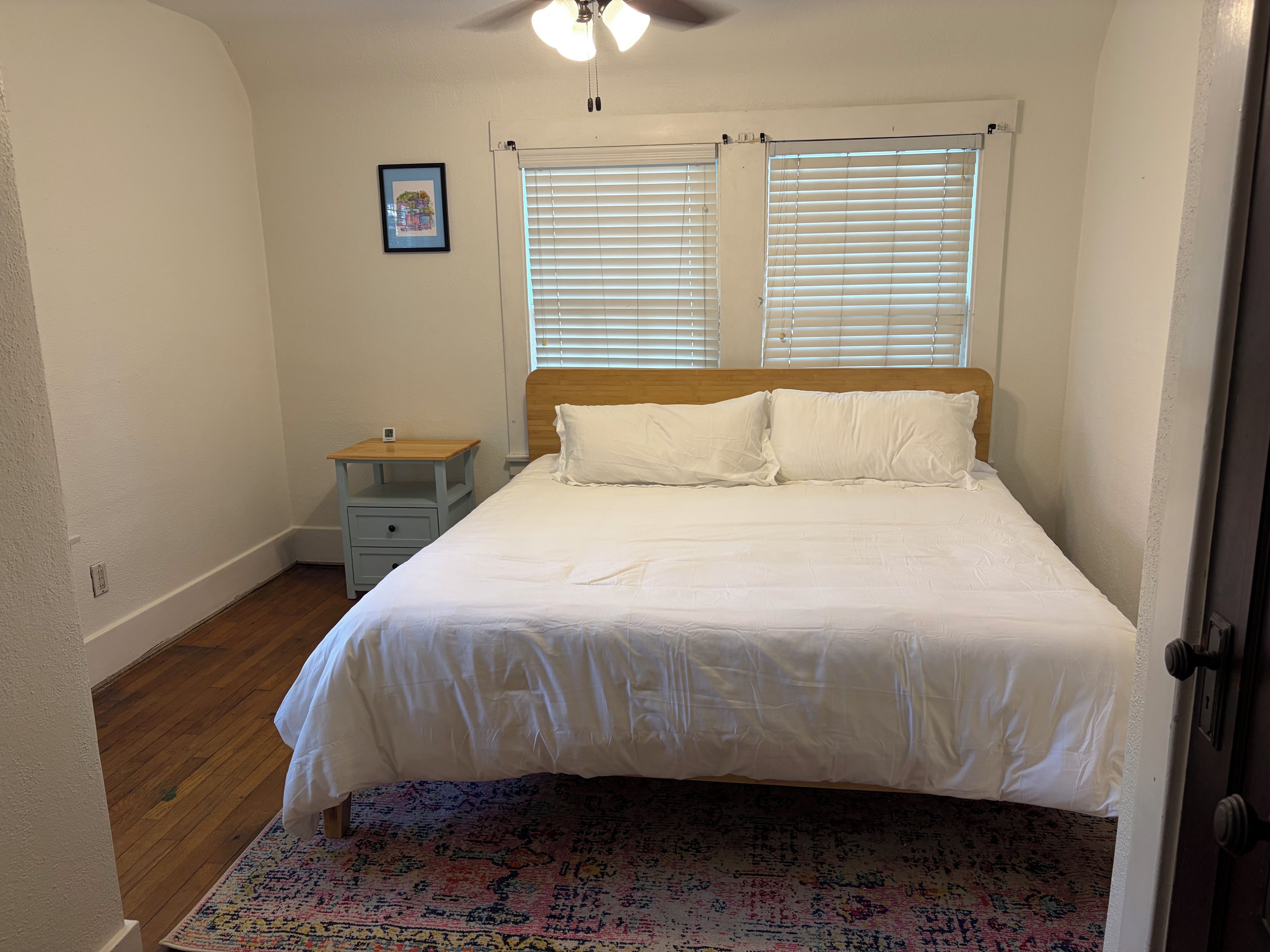 Quarto com cama, mesa de cabeceira, cabeceira de madeira, janela com persianas, tapete estampado e ventilador de teto.
