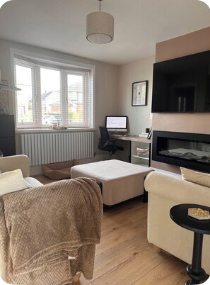 A living room with a sofa, ottoman, and a fireplace. There is a desk with a computer and a TV mounted on the wall.