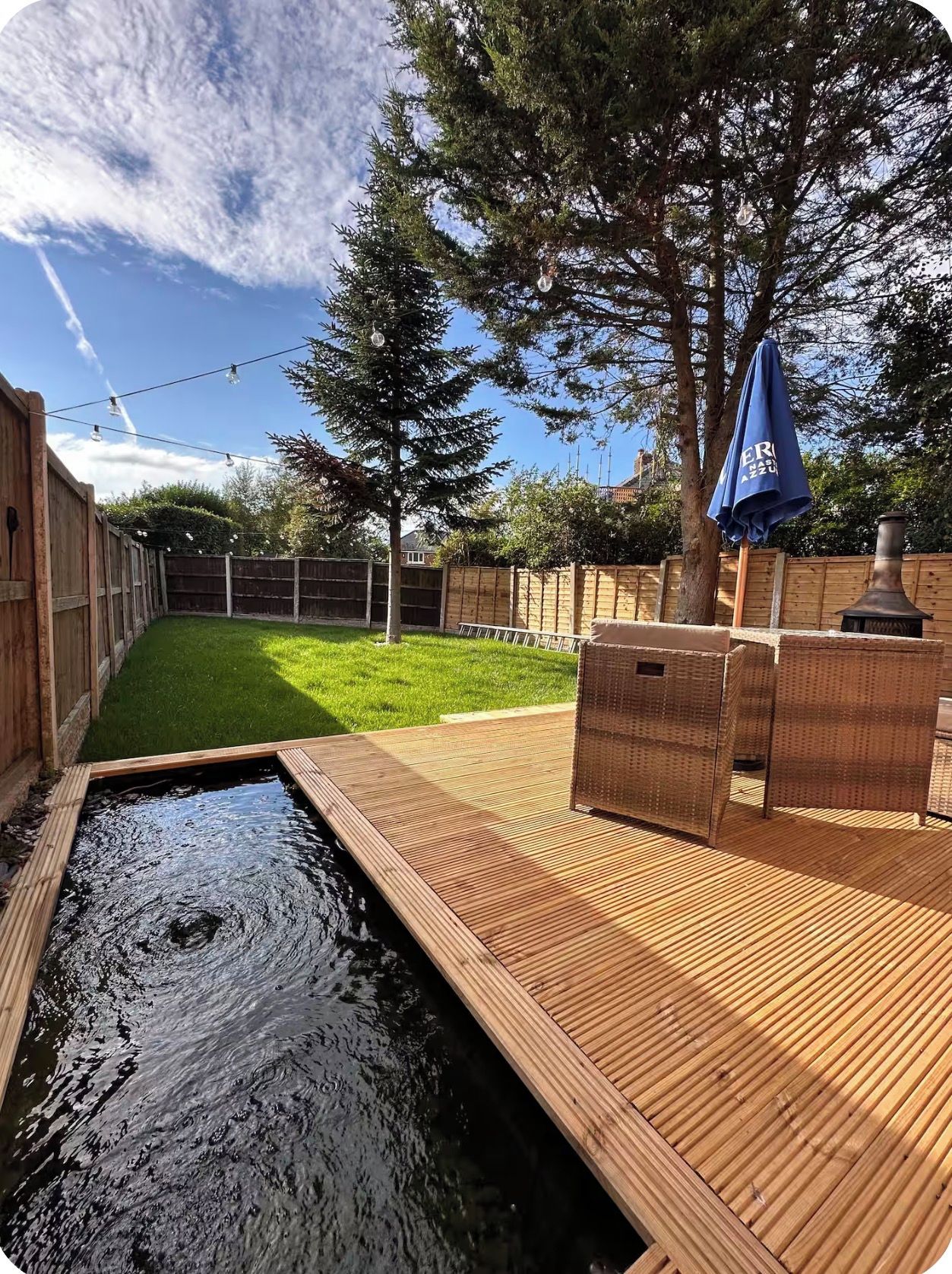 A backyard with a wooden deck, a small pond, a barbecue grill, and a pergola with a blue umbrella.