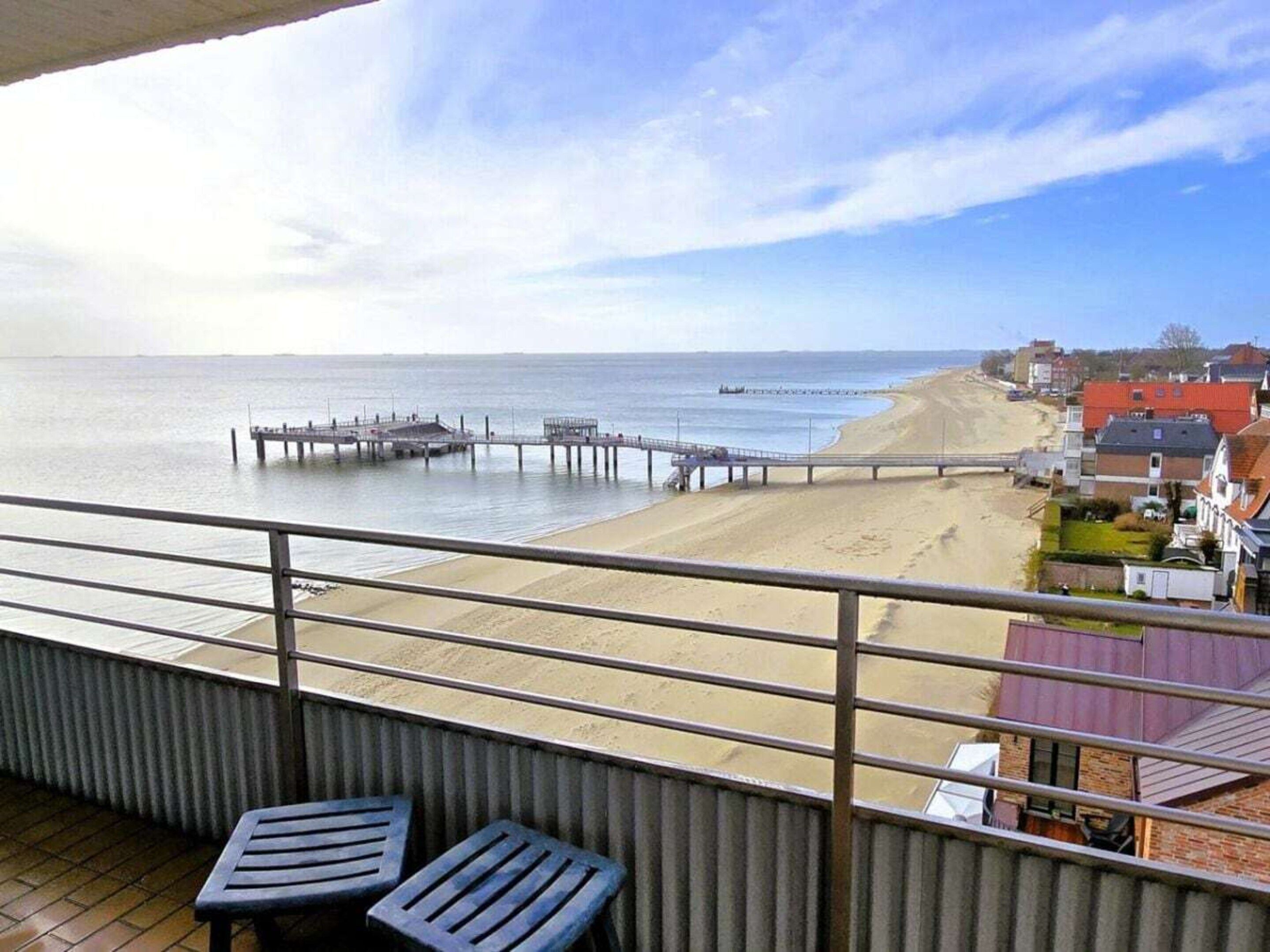 Uma varanda com duas cadeiras, vista para uma praia, um píer e uma fileira de casas.
