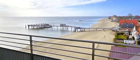 Uma varanda com duas cadeiras, vista para uma praia, um píer e uma fileira de casas.