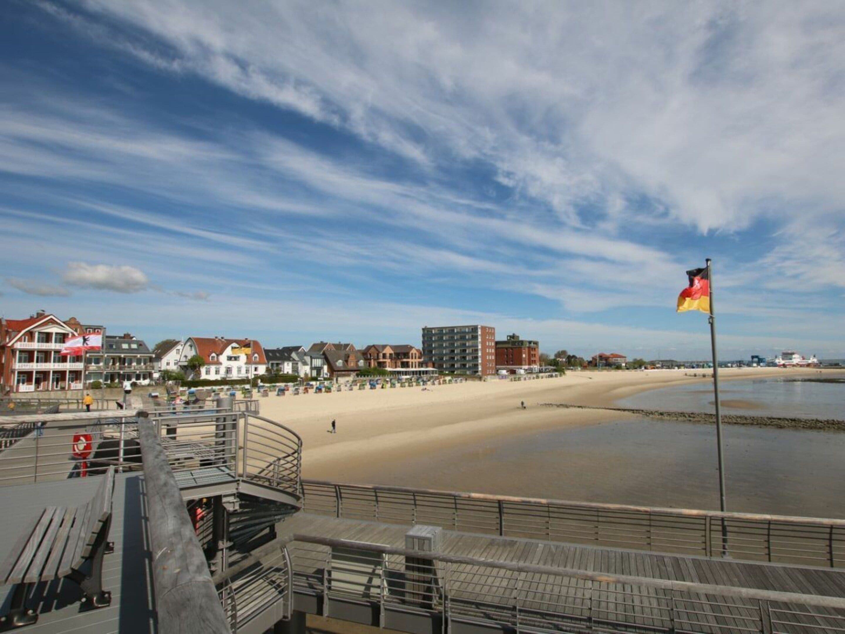 A coastal promenade with a sandy beach, a pier with a flag, and buildings in the background.