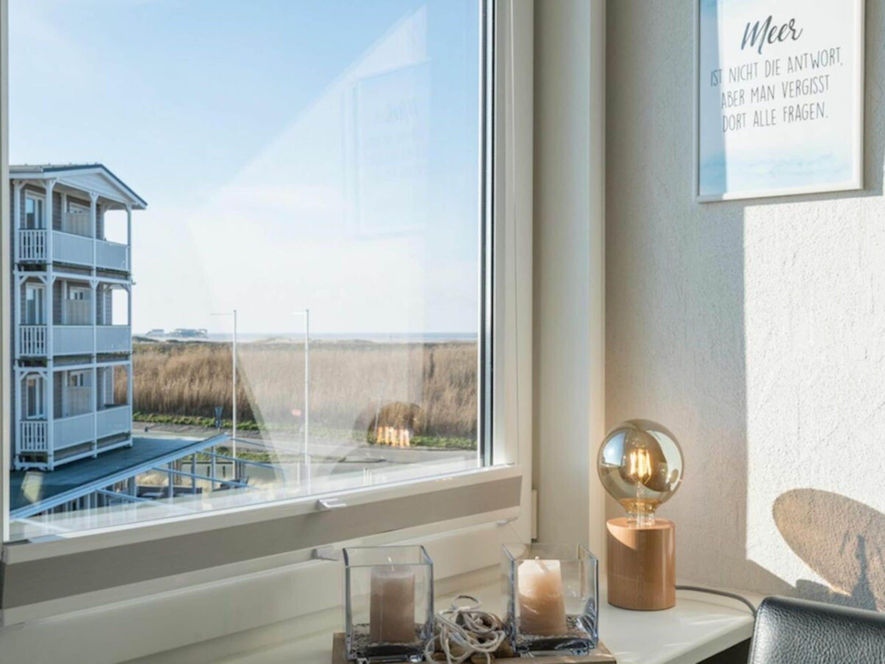 A room with a large window overlooking a building and a beach. There is a framed quote on the wall and a table with a candle and a lamp.