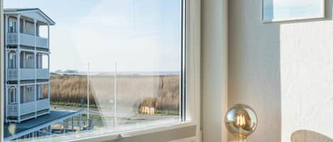 Ein Zimmer mit großem Fenster, das auf ein Gebäude und einen Strand blickt. An der Wand hängt ein gerahmter Spruch, und ein Tisch steht mit einer Kerze und einer Lampe darauf.