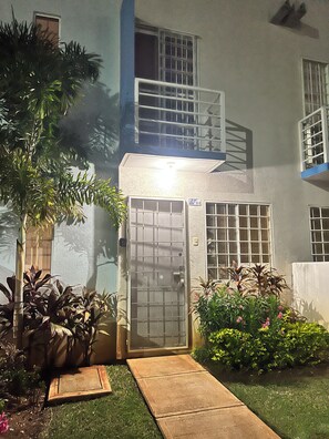 Edifício residencial com portão fechado, pequena varanda e jardim bem cuidado com diversas plantas e flores.