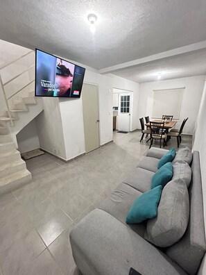 Ein modernes Wohnzimmer mit einem grauen Sofa, türkisfarbenen Kissen, einem Flachbildfernseher, einer Treppe und einem Essbereich mit einem Holztisch und Stühlen.