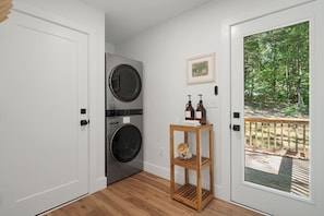 A laundry room with two stacked washing machines, a wooden stool with bottles, and a door leading to an outdoor deck with a view of trees. - Close to Blue Ridge Parkway, Mtn Views, Firepit (Asheville)