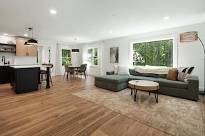A modern living space with a kitchen, dining area, and seating area, featuring hardwood floors, a sectional sofa, and large windows. - Close to Blue Ridge Parkway, Mtn Views, Firepit (Asheville)