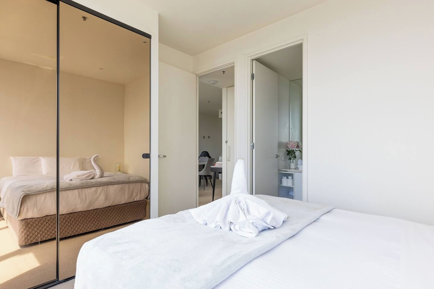 A hotel room with a bed, a glass partition, a bathroom with a mirror, and a small table with a chair.