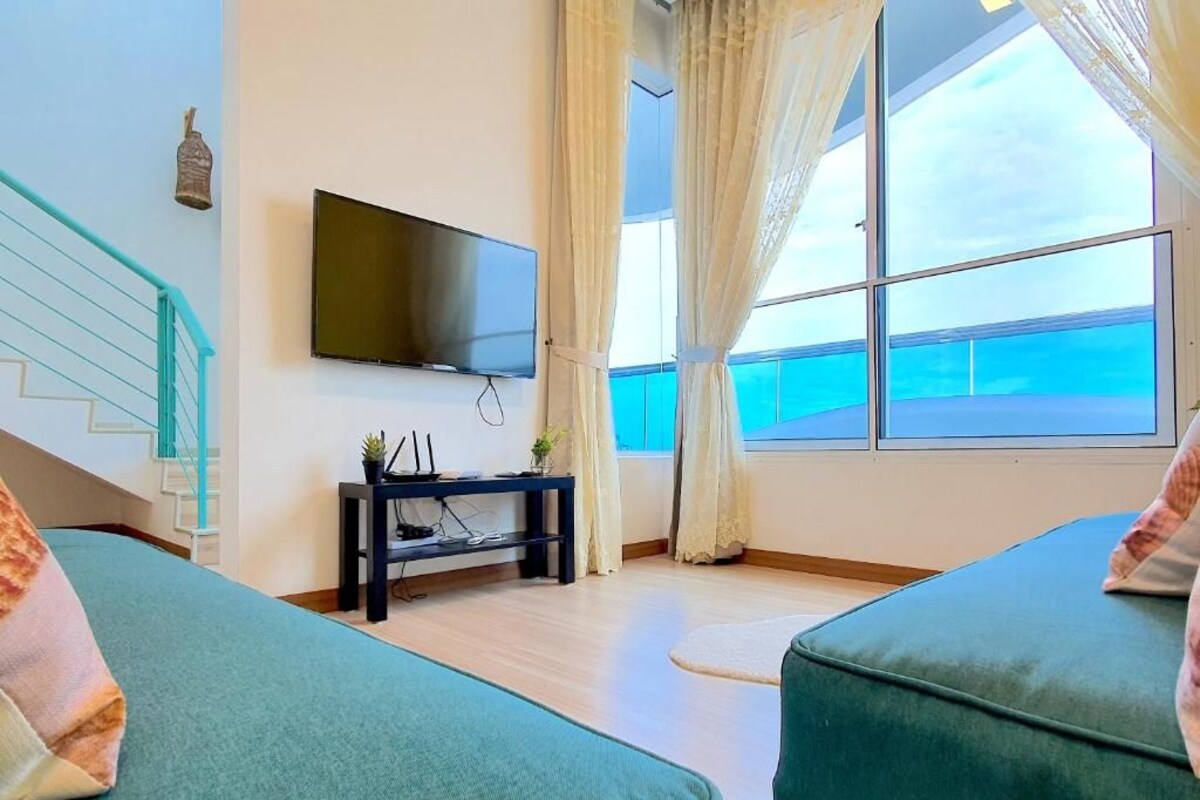 A room with a bed, a TV mounted on the wall, a small table with a plant, and a window with curtains.