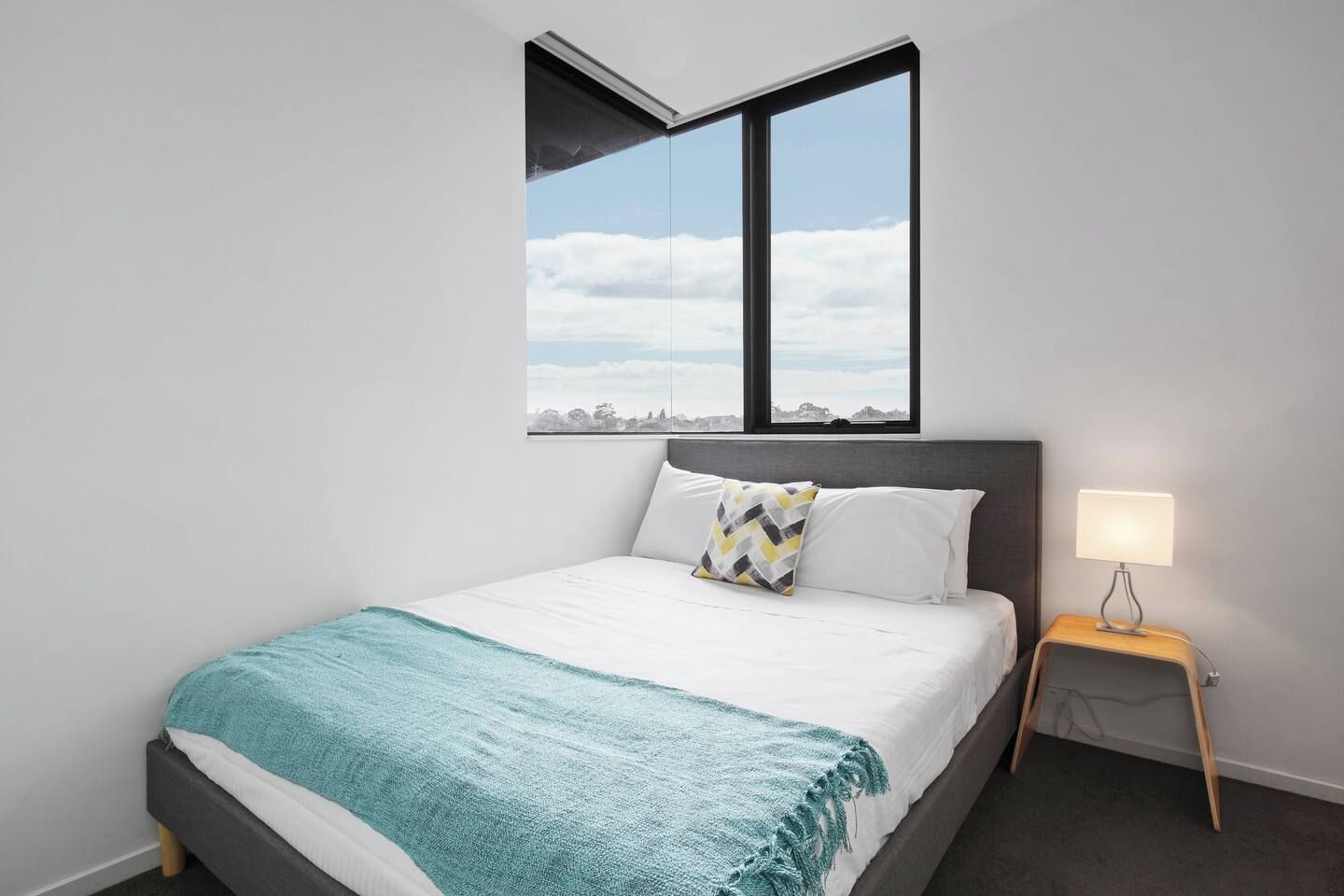 Een slaapkamer met een bed, nachtkastje, lamp en een raam met uitzicht op de lucht en wolken.