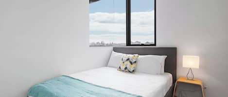 A bedroom with a bed, a nightstand, a lamp, and a window with a view of the sky and clouds.