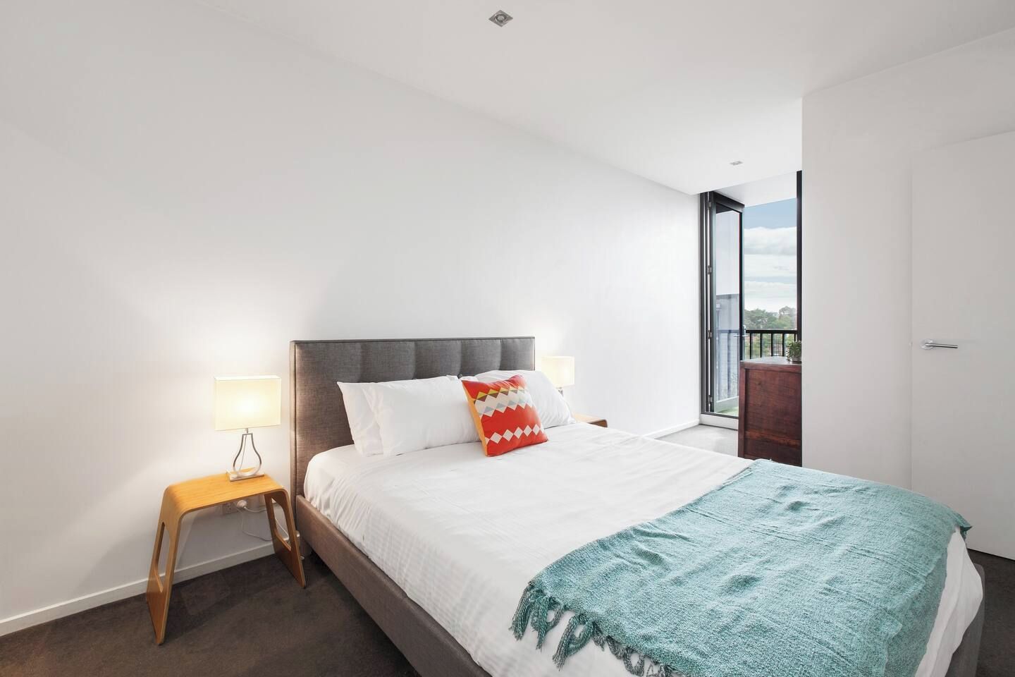 Een moderne slaapkamer met een bed, nachtlampjes, een klein houten nachtkastje en uitzicht door een groot raam.