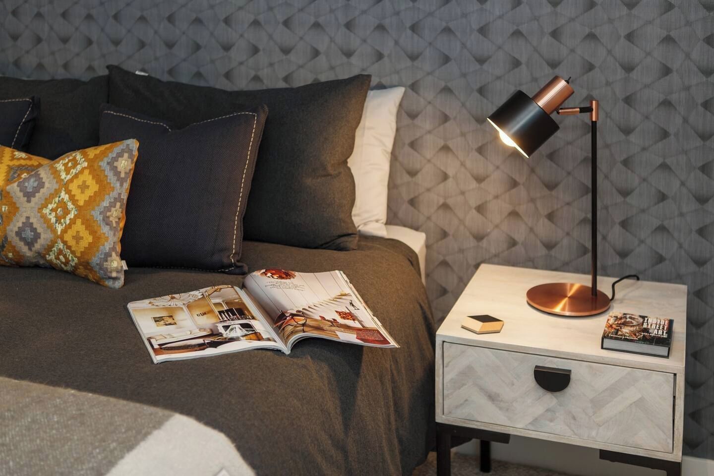 Een keurig opgemaakt bed met een tijdschrift, een lamp en een boek op een nachtkastje.