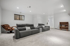 Ein modernes Wohnzimmer mit einem grauen Ecksofa, einem braunen Ledersessel, einem Holzregal und einem Fenster mit Jalousien.