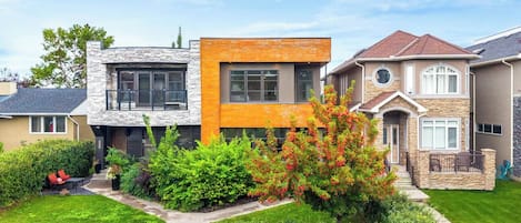 A modern two-story house with a stone facade, a wooden front section, and a well-maintained garden with various shrubs and a tree.