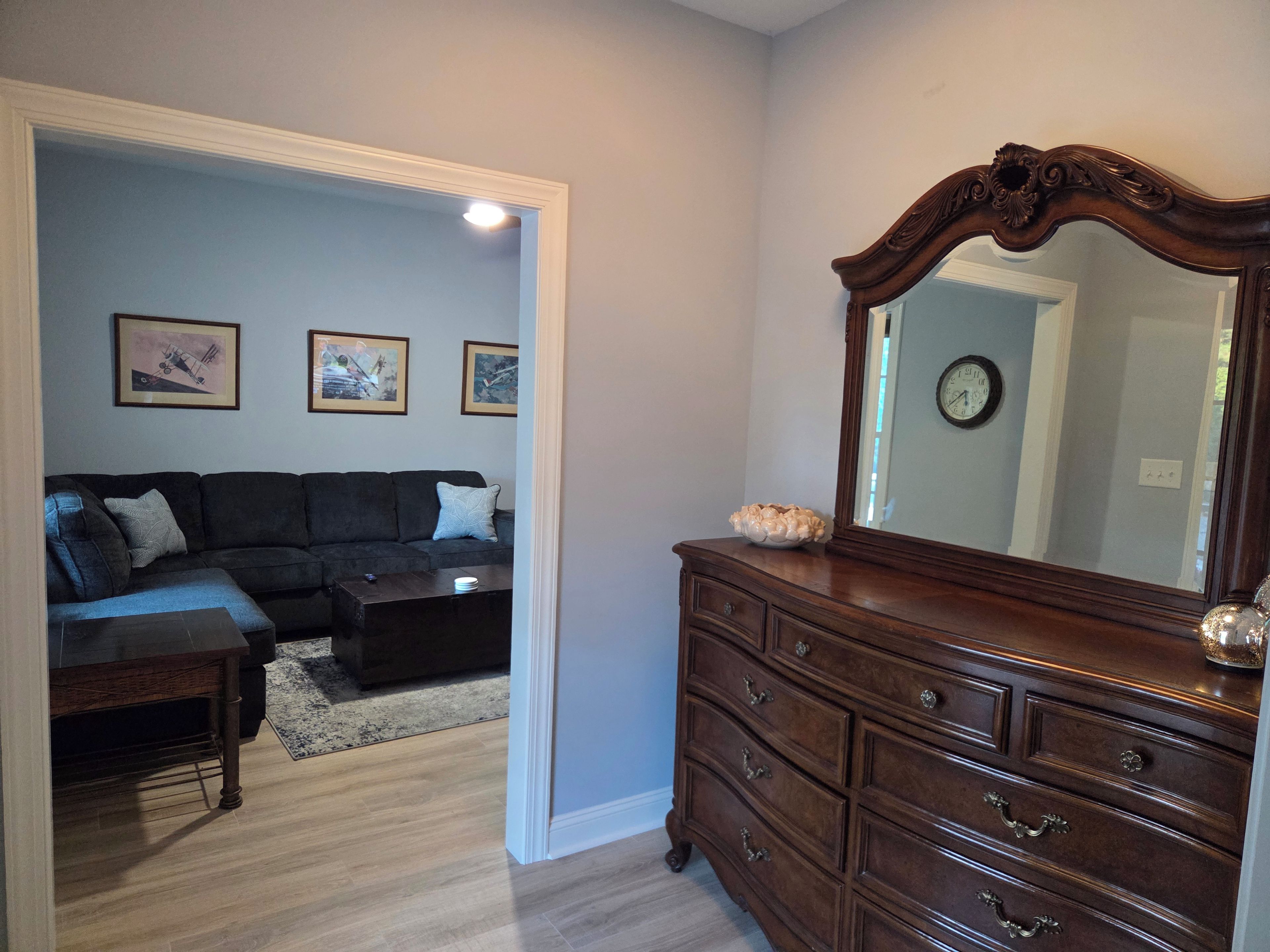 A living room with a dark-colored sofa, a coffee table, and a wooden dresser with a mirror. There are framed pictures on the wall and a clock.