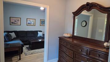 A living room with a dark-colored sofa, a coffee table, and a wooden dresser with a mirror. There are framed pictures on the wall and a clock.
